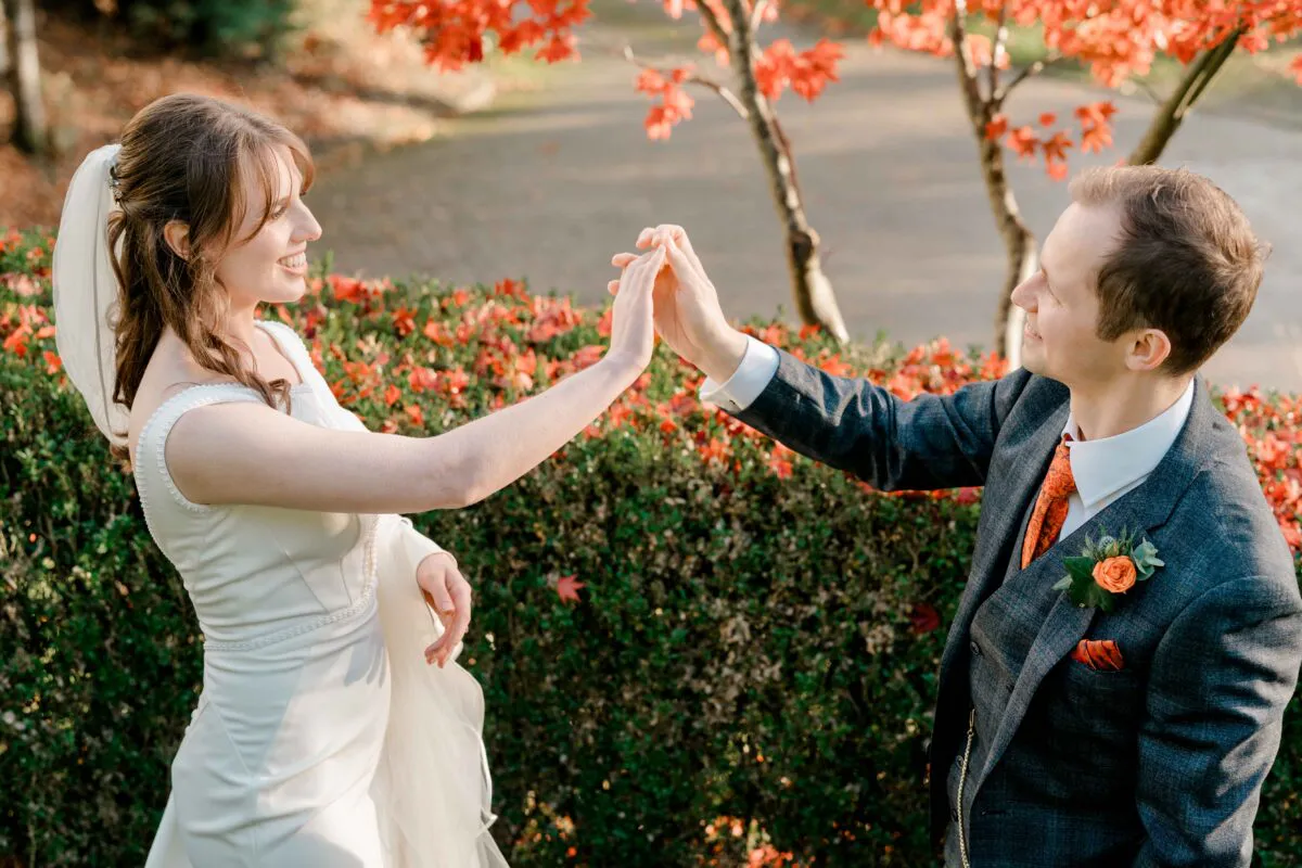 A newlywed couple laugh and hold hands outdoors against a backdrop of autumn foliage and vibrant red leaves during an afternoon portrait session.