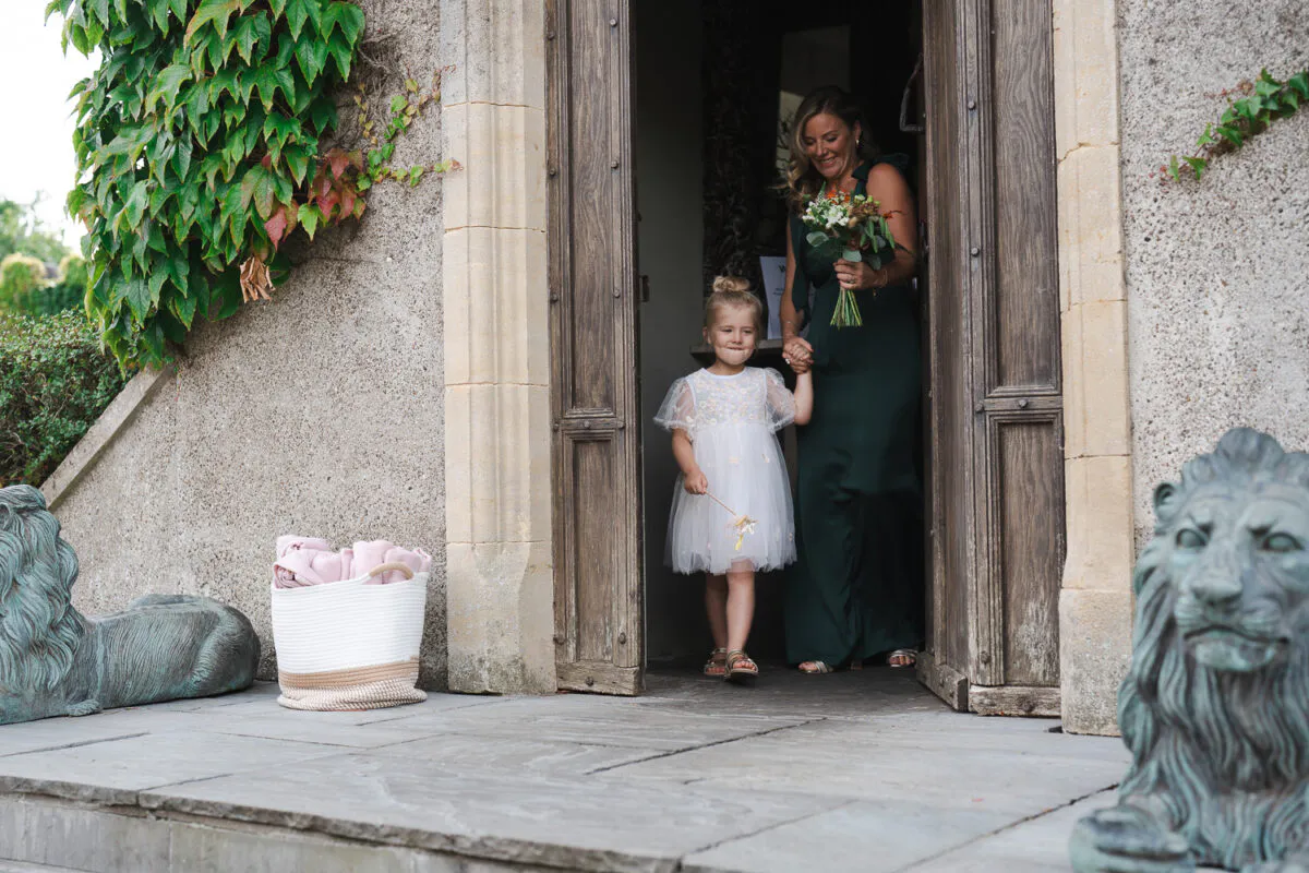 A young flower girl in a white dress and a bridesmaid in a dark green gown walk hand-in-hand through a stone doorway holding a bouquet.