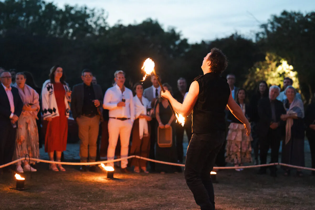 A fire performer entertains an outdoor evening crowd of wedding guests gathered on grass at dusk.
