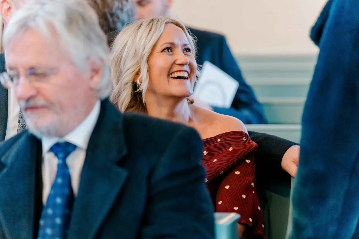 A blonde female wedding guest wearing an off-the-shoulder burgundy dress laughs joyfully while seated in a church pew.