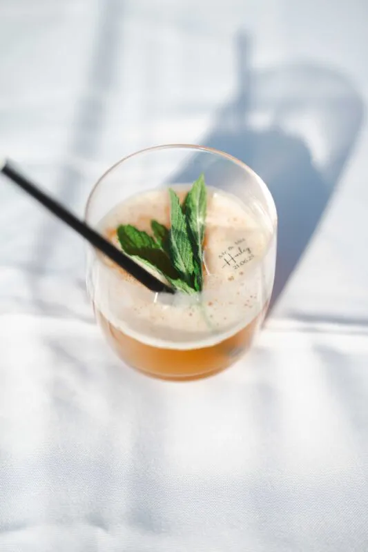 A refreshing signature cocktail garnished with fresh mint and served in a personalised glass sits on a bright, sunlit white surface during a wedding drinks reception.