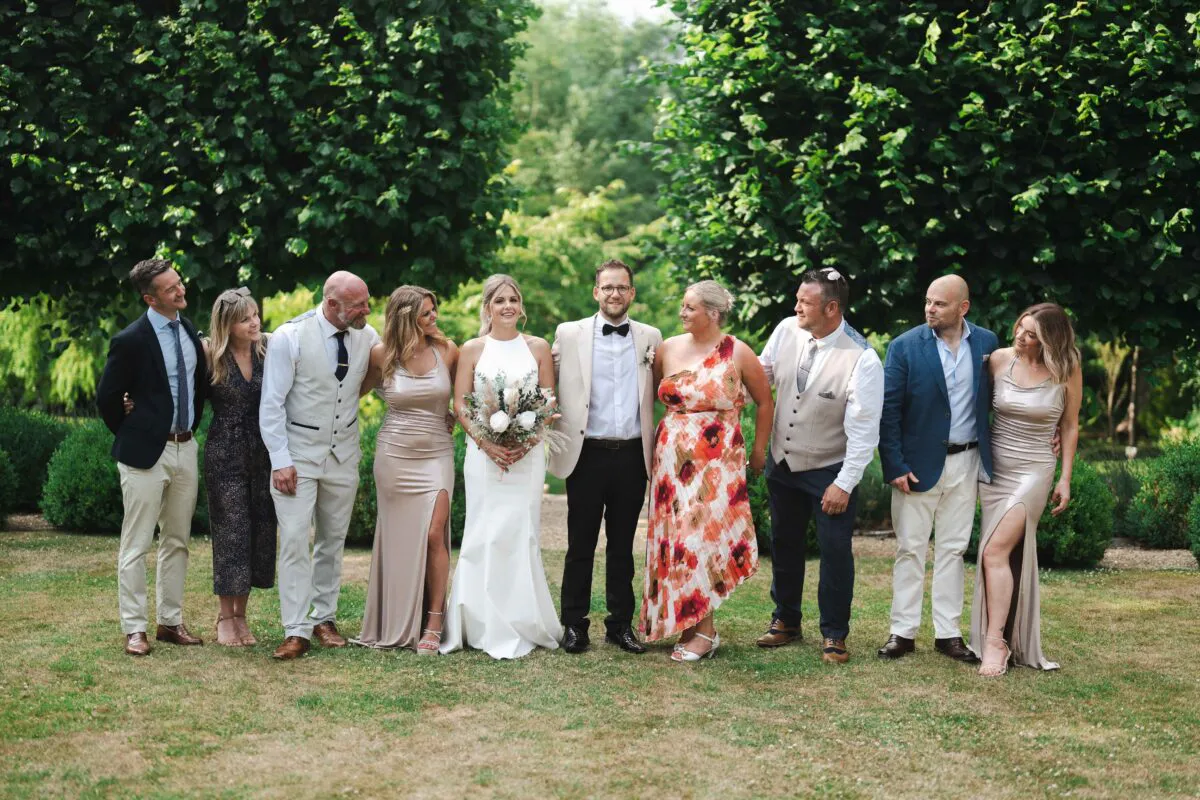 The bride and groom pose happily alongside their wedding party in a lush green garden setting surrounded by tall trees.
