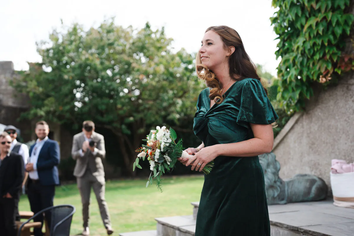A smiling bridesmaid wearing a dark green velvet dress holding a bouquet stands outside near stone steps while guests chat in the background.