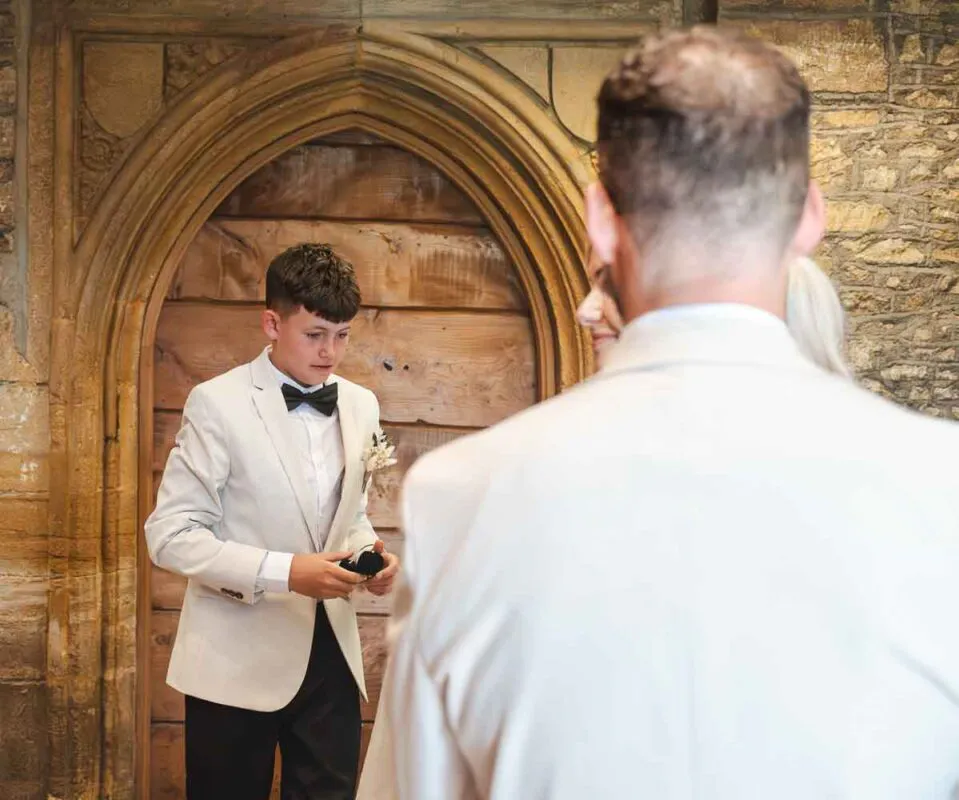 A young page boy wearing a white tuxedo jacket and black bow tie stands in front of a historic stone archway and wooden door, holding a ring box, viewed over the shoulder of another person.