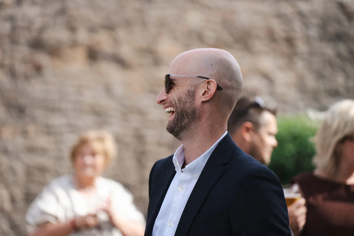 A bald wedding guest wearing sunglasses and a dark suit jacket laughs heartily outdoors against a rustic stone wall.