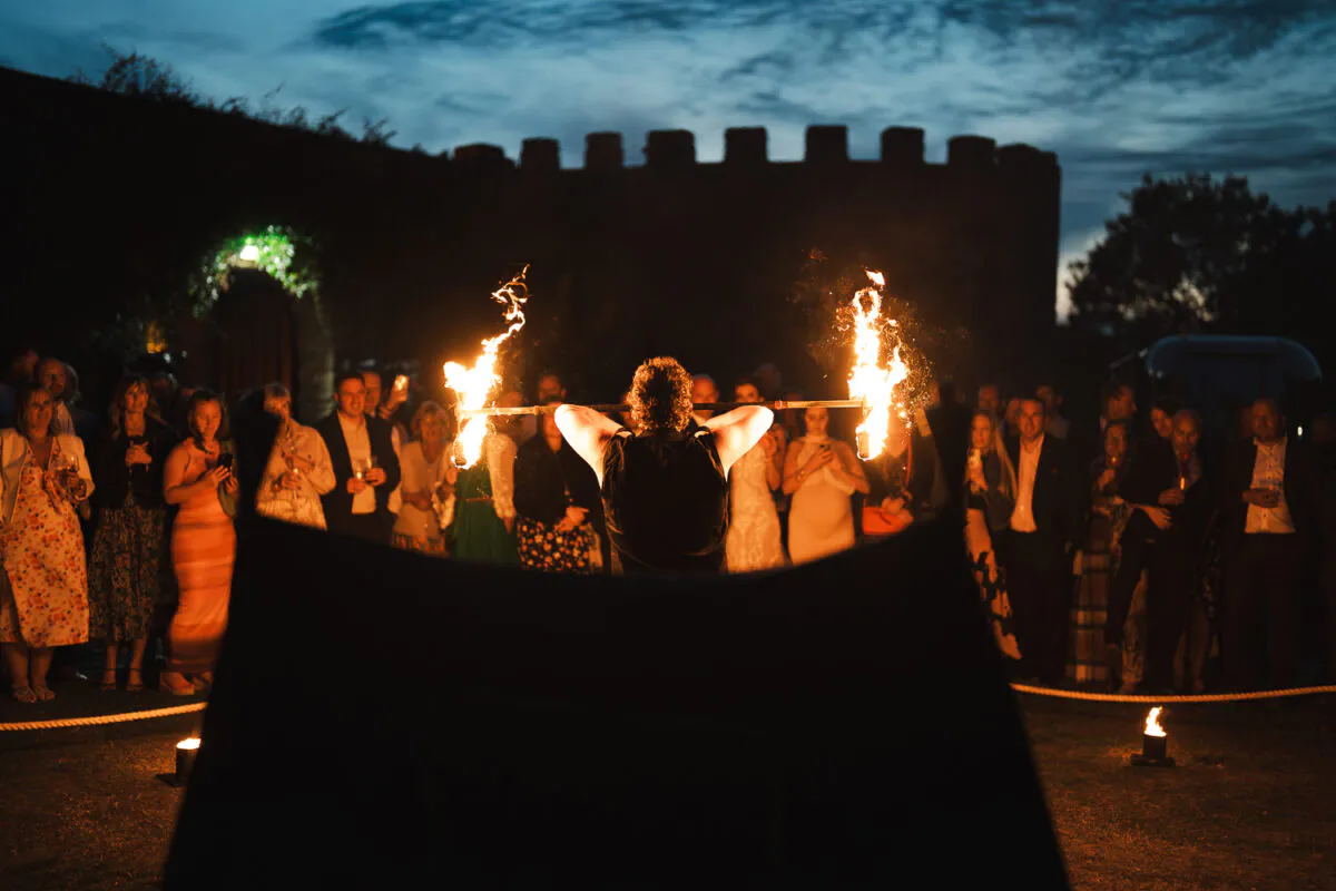A fire performer holds a blazing staff at an outdoor evening reception with a castle backdrop while guests watch on.