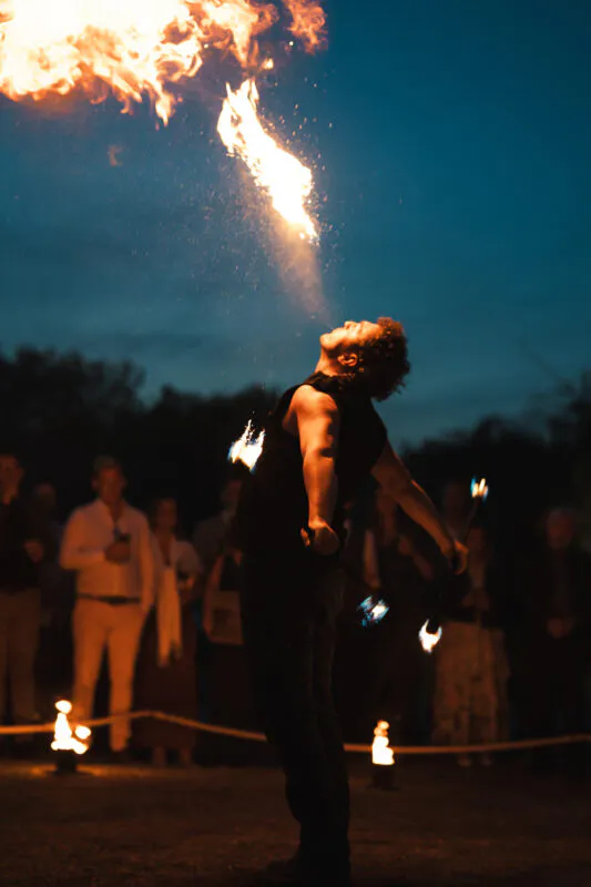 A skilled fire performer entertains wedding guests with a dramatic fire breathing stunt during an evening reception under the night sky.