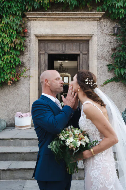 A newlywed couple share a playful kissing moment outside a stone doorway framed by lush green ivy, with the bride holding a textured bouquet of white flowers and greenery.