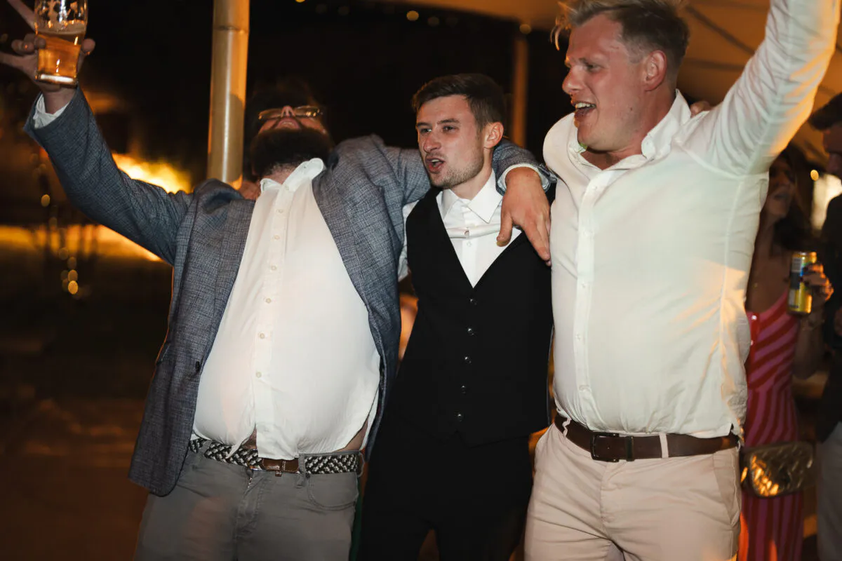 Three wedding guests celebrate joyfully on the dance floor with arms raised in the air during the evening reception.