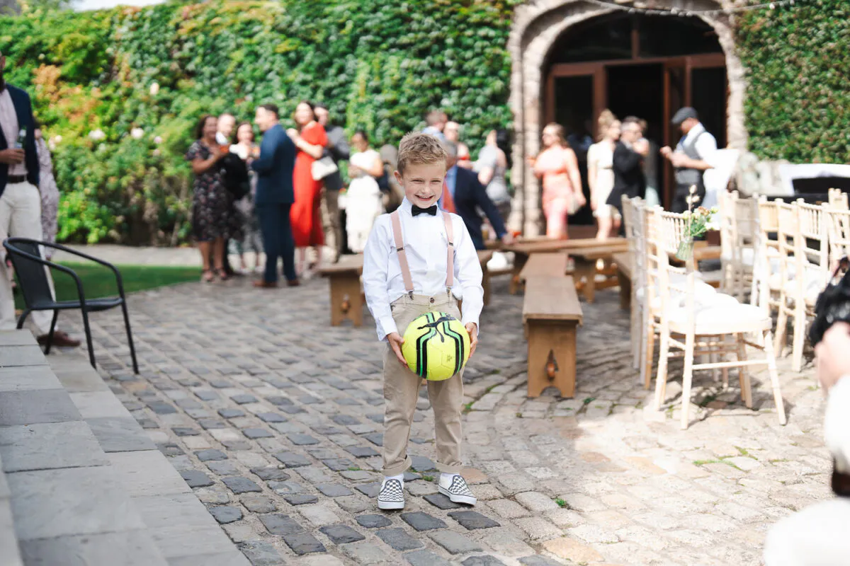 A young page boy in a white shirt, beige trousers, and trainers smiling and holding a football in a cobbled courtyard during an outdoor wedding.
