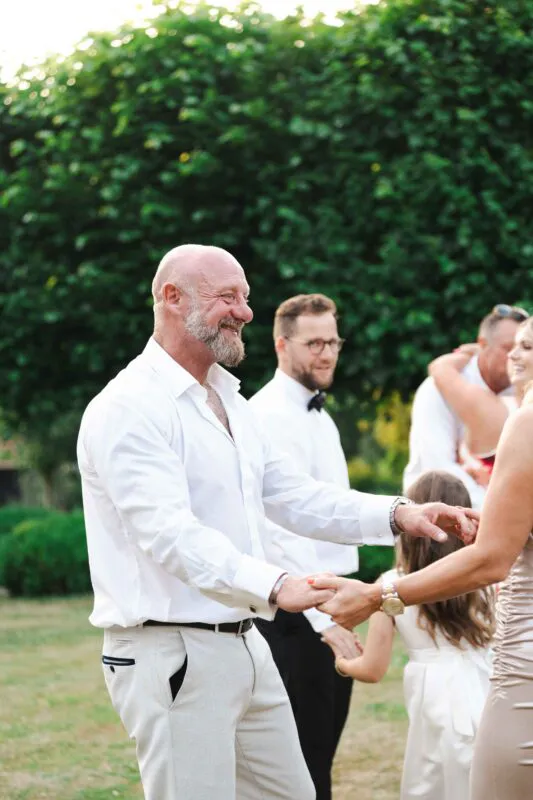A smiling, bearded older man in a white shirt and trousers holds hands and dances with a guest in an outdoor garden reception setting.