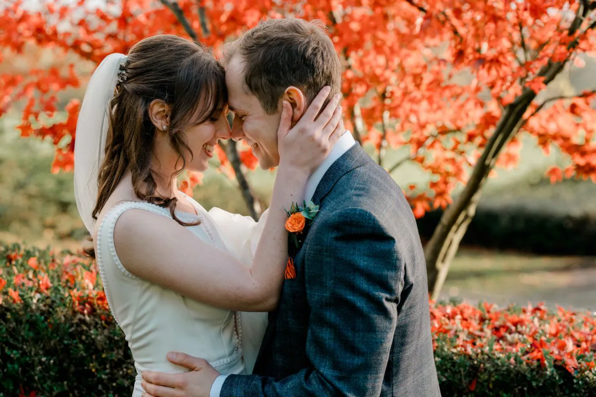 Newlywed bride and groom stand close together nose to nose outdoors amidst vibrant orange autumn foliage during golden hour.