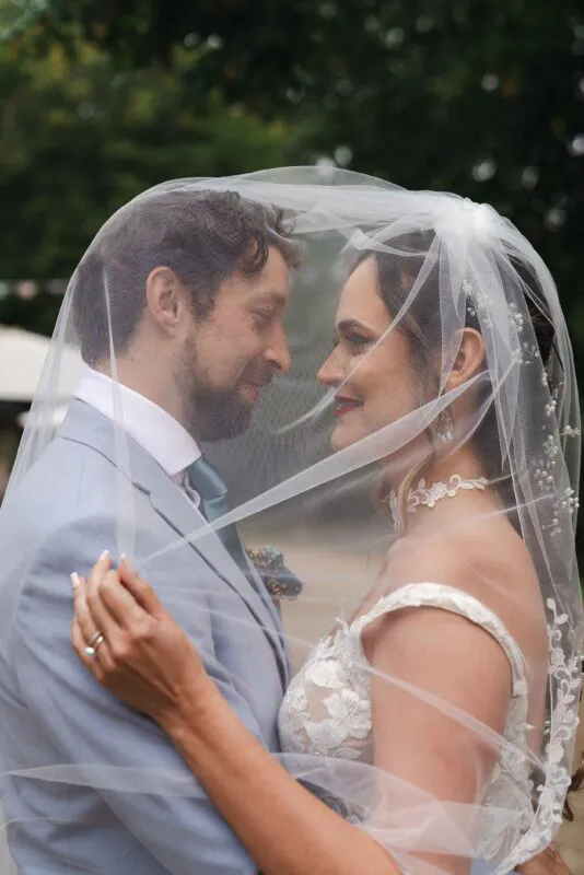 A newlywed couple share a tender moment standing nose to nose beneath a sheer embellished bridal veil.