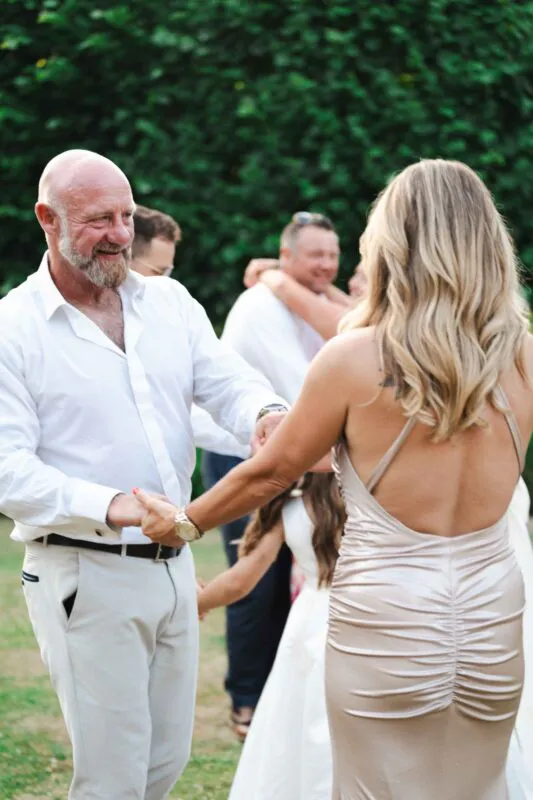 A mature couple joyfully dance together outdoors, with the smiling groom in a white shirt and light trousers holding hands with the bride in a golden satin backless gown.