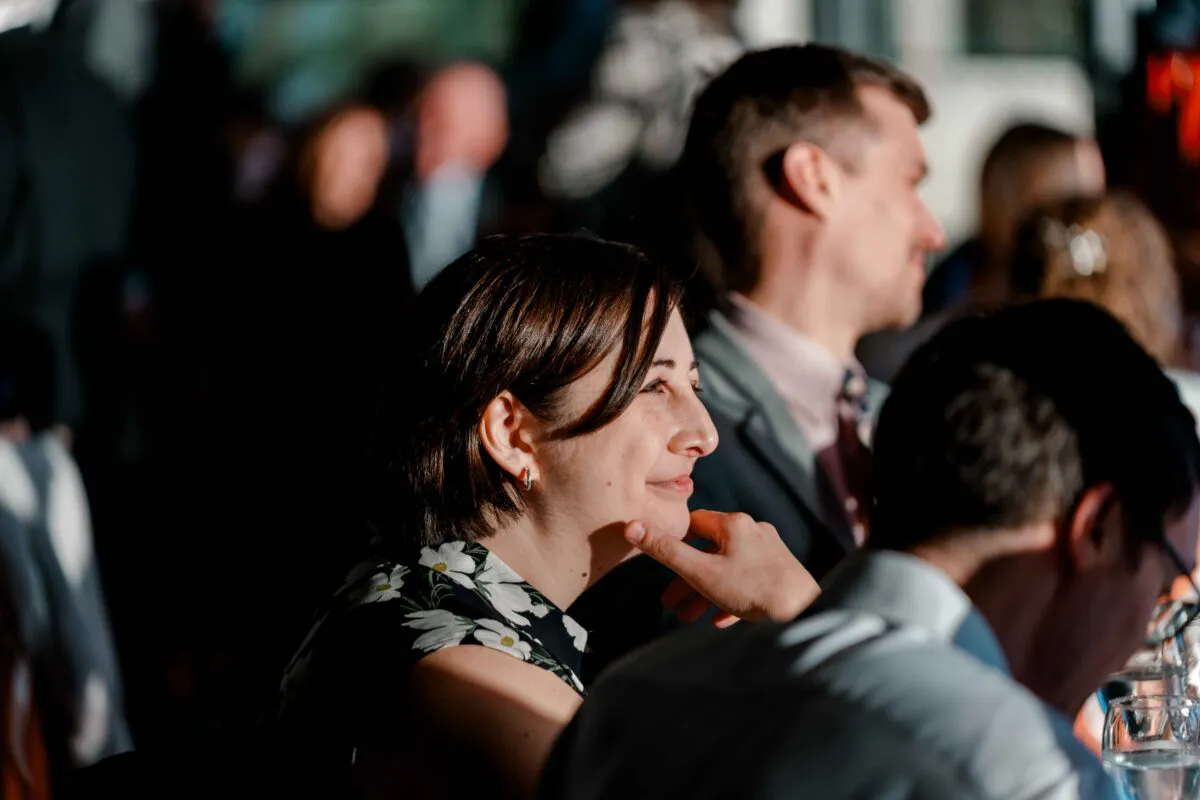 A guest smiles thoughtfully during the wedding breakfast, illuminated by warm side light with other guests blurred in the background.