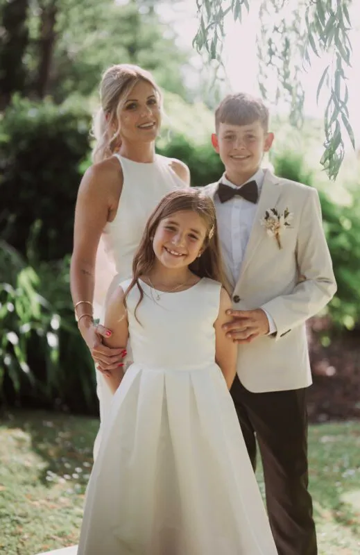 An outdoor family group portrait featuring a bride in a white halterneck dress standing alongside a smiling flower girl in a white dress and a page boy in a cream suit with a bow tie.