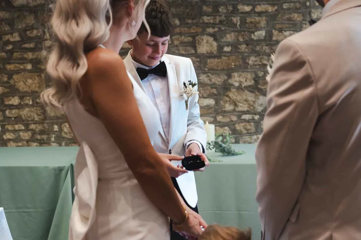 A young page boy wearing a white tuxedo jacket and black bow tie proudly holds out an open ring box during an indoor wedding ceremony.