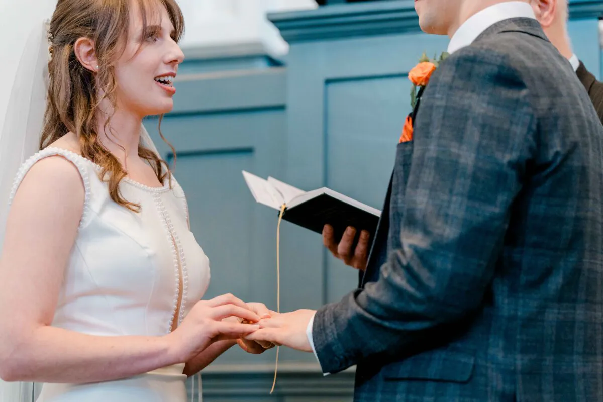 A smiling bride wearing a pearl-trimmed white dress exchanges wedding rings with the groom, who is dressed in a dark checked suit during their ceremony.