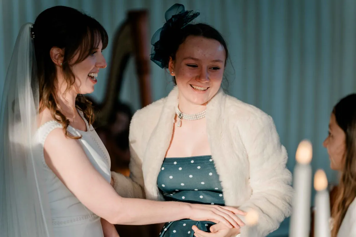A smiling bride wearing a veil and white dress shares a joyful moment with a guest in a polka dot dress, faux fur stole, and fascinator.