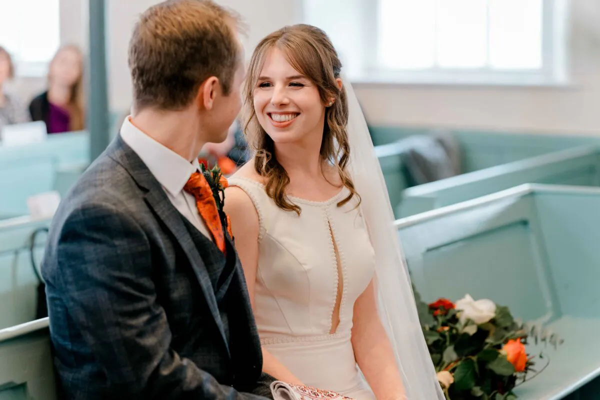 A smiling bride and groom gaze lovingly at each other while seated together in a church pew during their wedding ceremony.