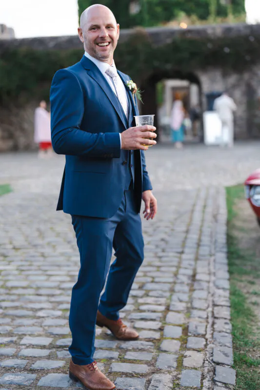 A smiling groom in a blue suit and brown brogues holding a drink while standing on a cobbled courtyard during a wedding reception.