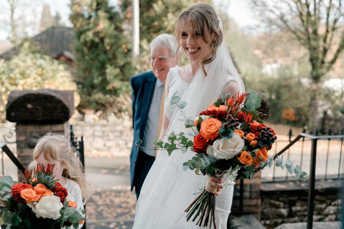 A smiling bride holding a vibrant orange and white bouquet walks down the aisle accompanied by her father, while a young flower girl holds a matching bouquet in the foreground.