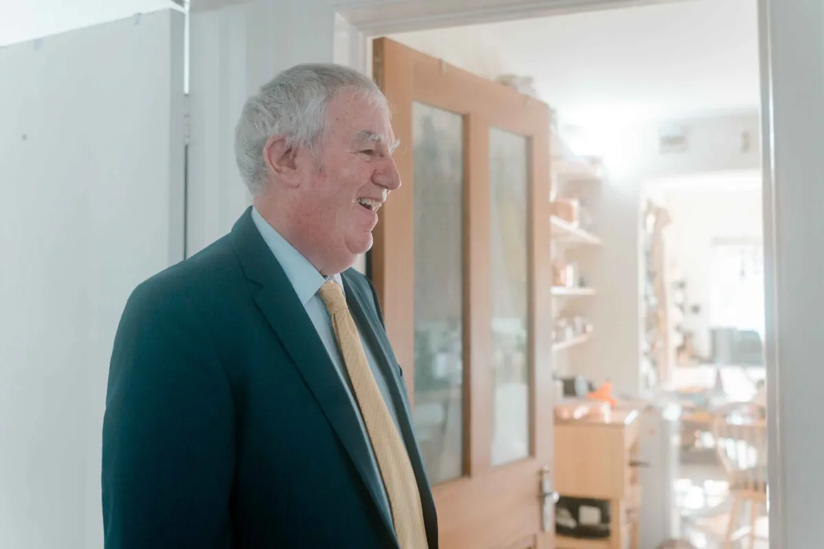 An elderly gentleman in a dark suit and yellow tie smiles happily while standing near an open wooden doorway.
