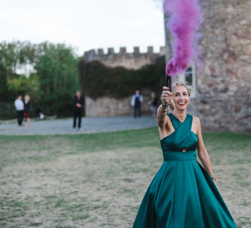 A smiling wedding guest in an emerald green satin halterneck gown holds up a vibrant pink smoke bomb outside a historic stone castle.