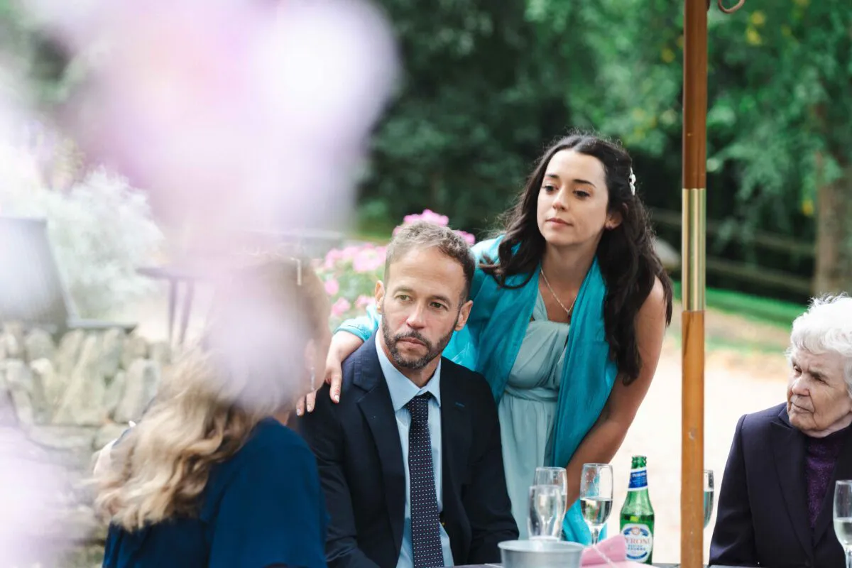 Wedding guests mingling and conversing outdoors during a reception, captured naturally through soft foreground foliage.
