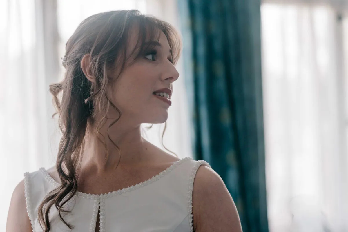 A beautiful bride looking over her shoulder with soft natural light illuminating her half-up hairstyle and beaded wedding dress during morning preparations.