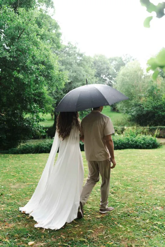 A newlywed couple walks hand in hand across a lush green lawn under a dark umbrella during a gentle rain shower.