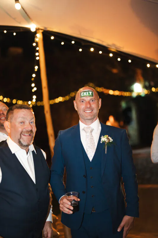 A smiling groom wearing a blue three-piece suit and a small exit sticker on his forehead stands with a guest at a nighttime marquee reception.