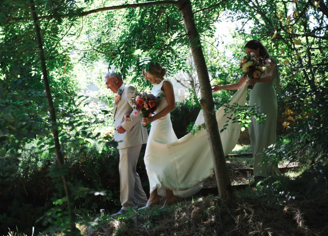 A smiling bride walking through a sunlit woodland accompanied by her father, with a bridesmaid carrying her long train behind them.
