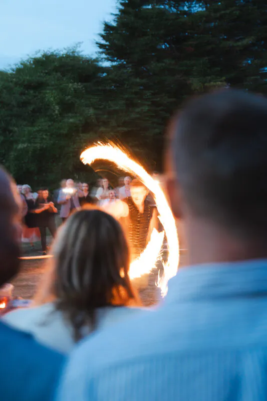 An exciting evening entertainment display features a fire performer twirling a flaming staff, viewed past the blurred shoulders of wedding guests watching in awe.