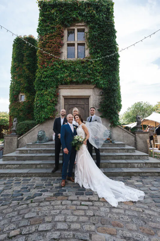 The bride and groom pose with wedding guests on a cobbled courtyard in front of a historic ivy-covered tower.
