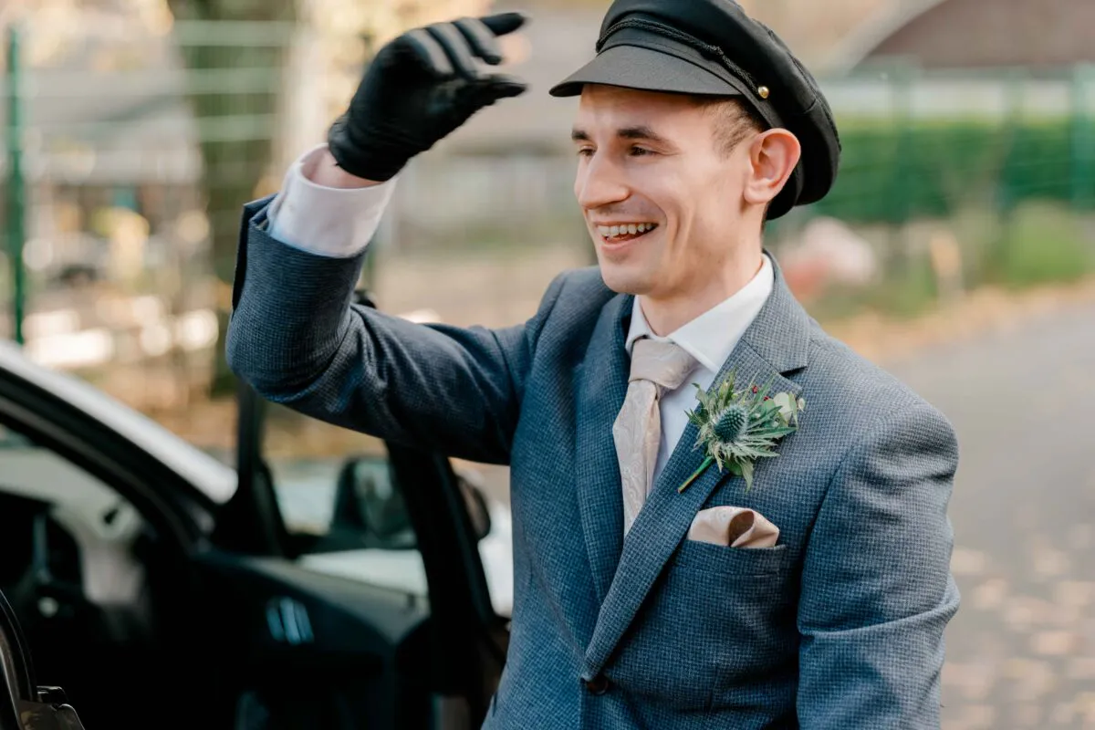 A smiling groom wearing a grey textured suit, patterned tie, and a thistle buttonhole raises a black-gloved hand near a car door.