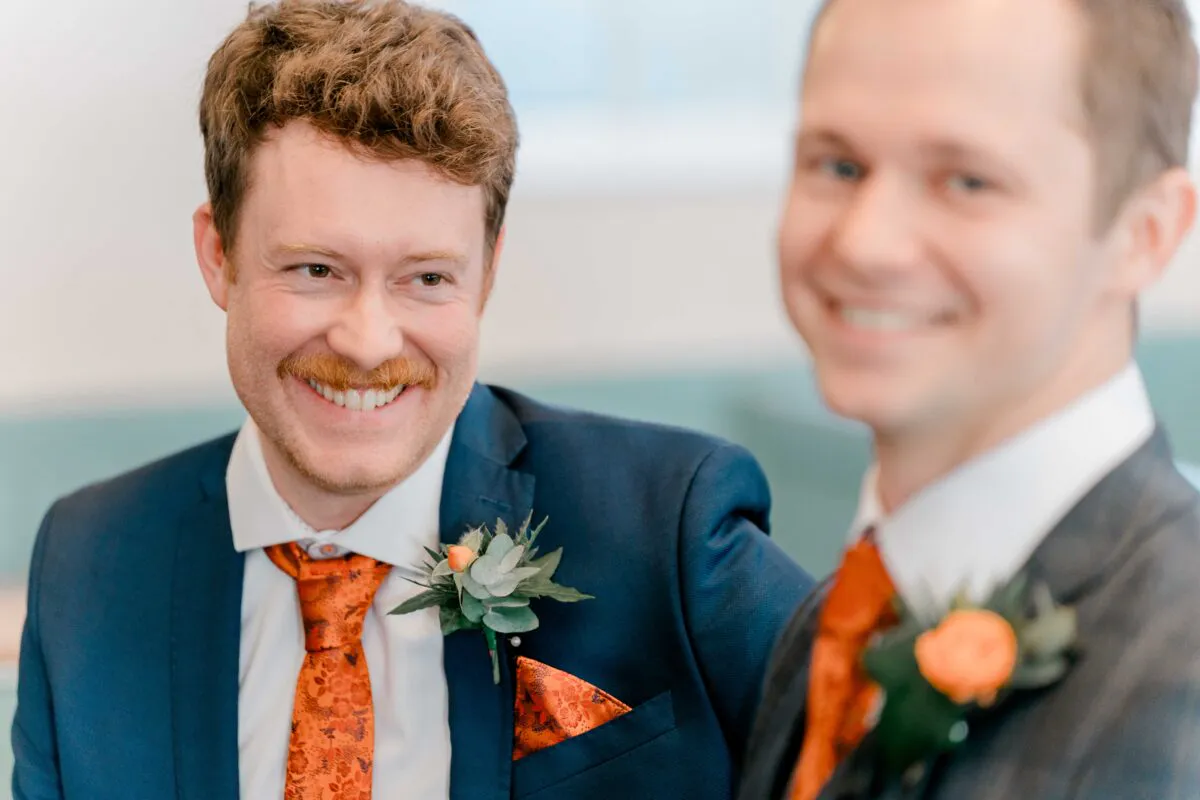 Two smiling grooms in navy suits and patterned orange ties share a happy moment during the wedding reception.