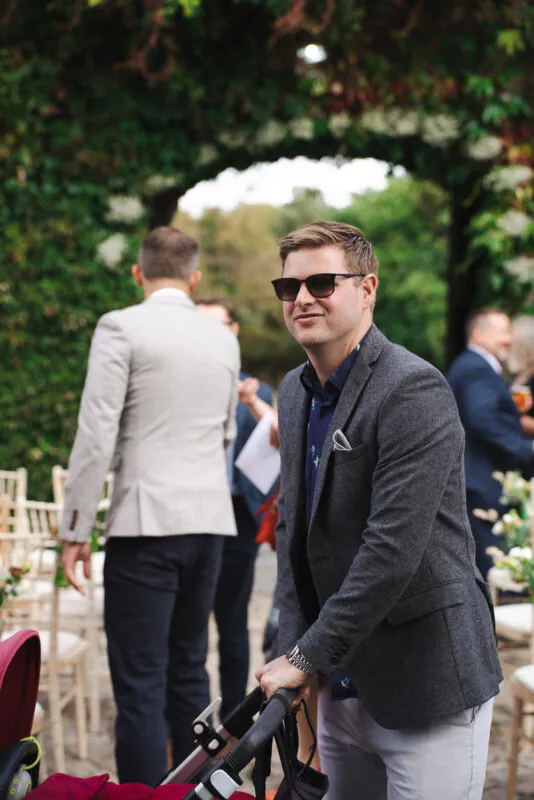 A wedding guest wearing a grey suit and sunglasses smiles while pushing a pram outdoors during a reception.
