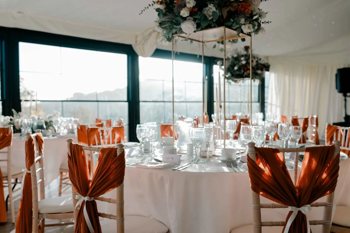 An elegantly styled wedding reception in a marquee featuring round tables adorned with white linens and striking burnt orange chair sashes.