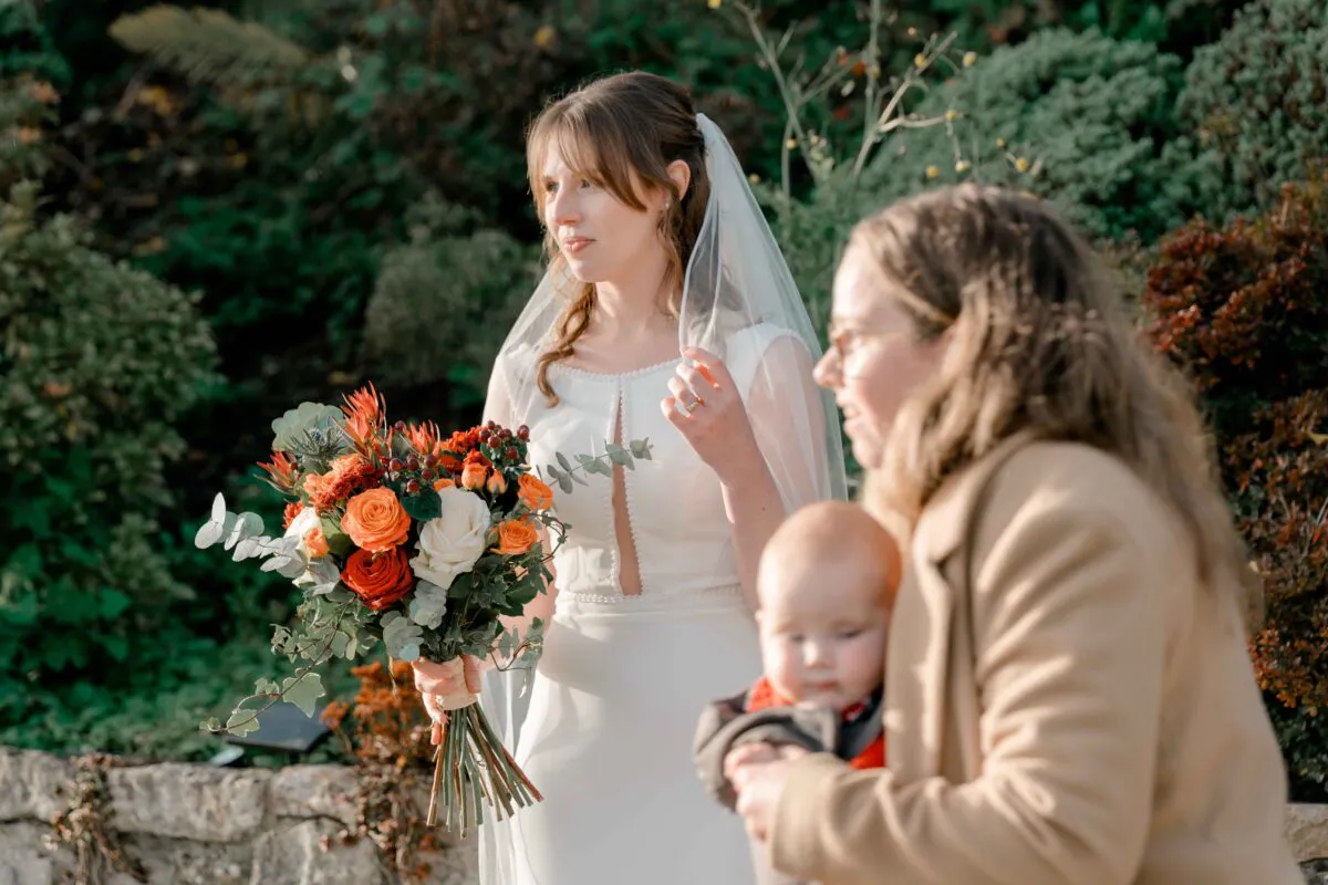 A bride holding a vibrant autumn bouquet featuring orange roses stands outdoors next to a guest holding a baby.