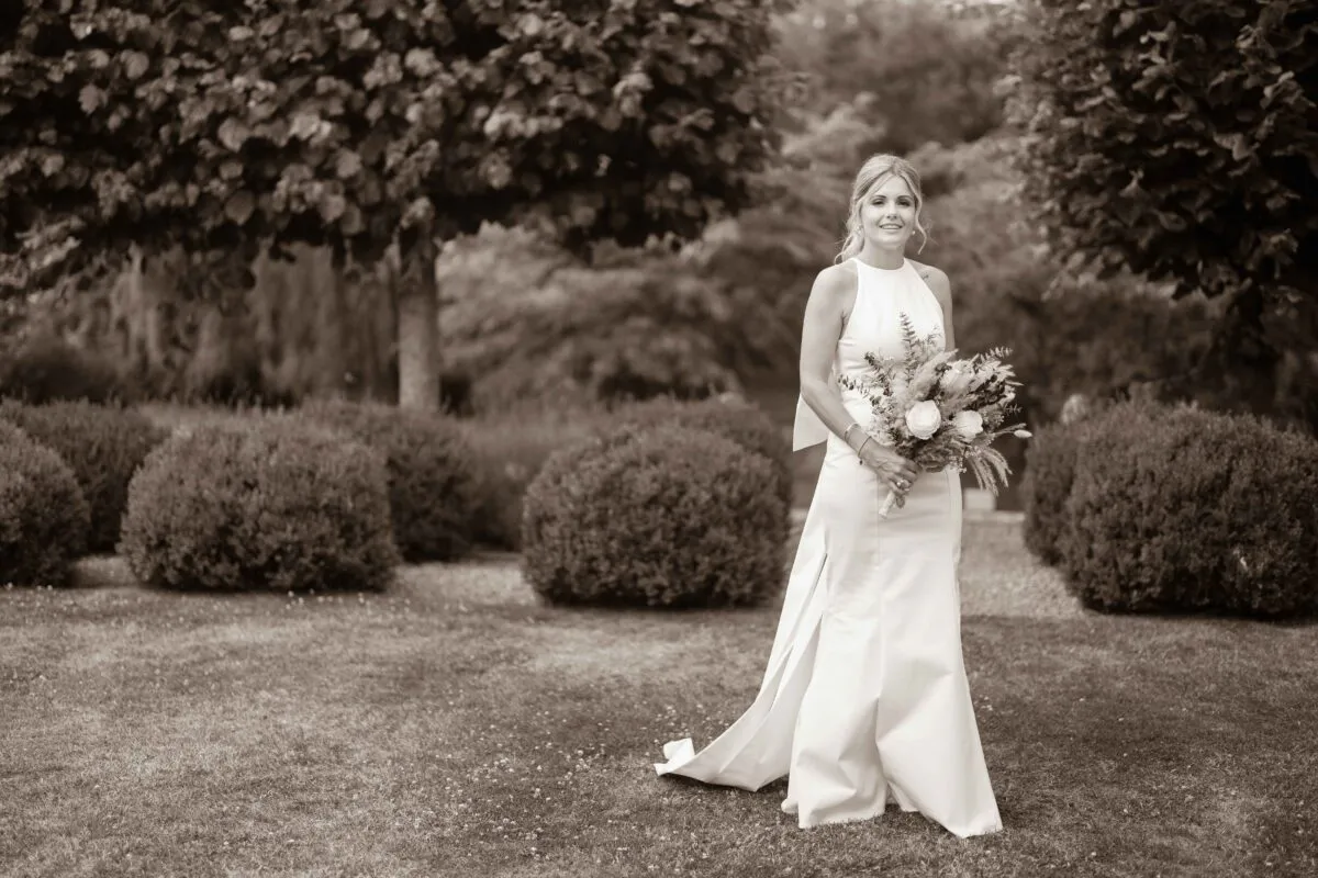 A stunning sepia-toned portrait of a smiling bride holding a textured bouquet in a formal garden with manicured topiary.