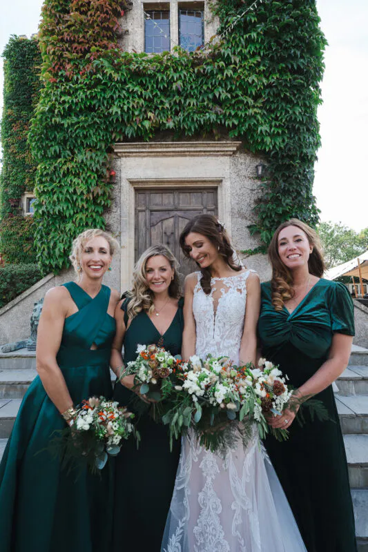 A stunning bride in an illusion lace wedding dress stands alongside her bridesmaids in rich emerald green gowns, holding hand-tied bouquets outside an ivy-covered stone building.