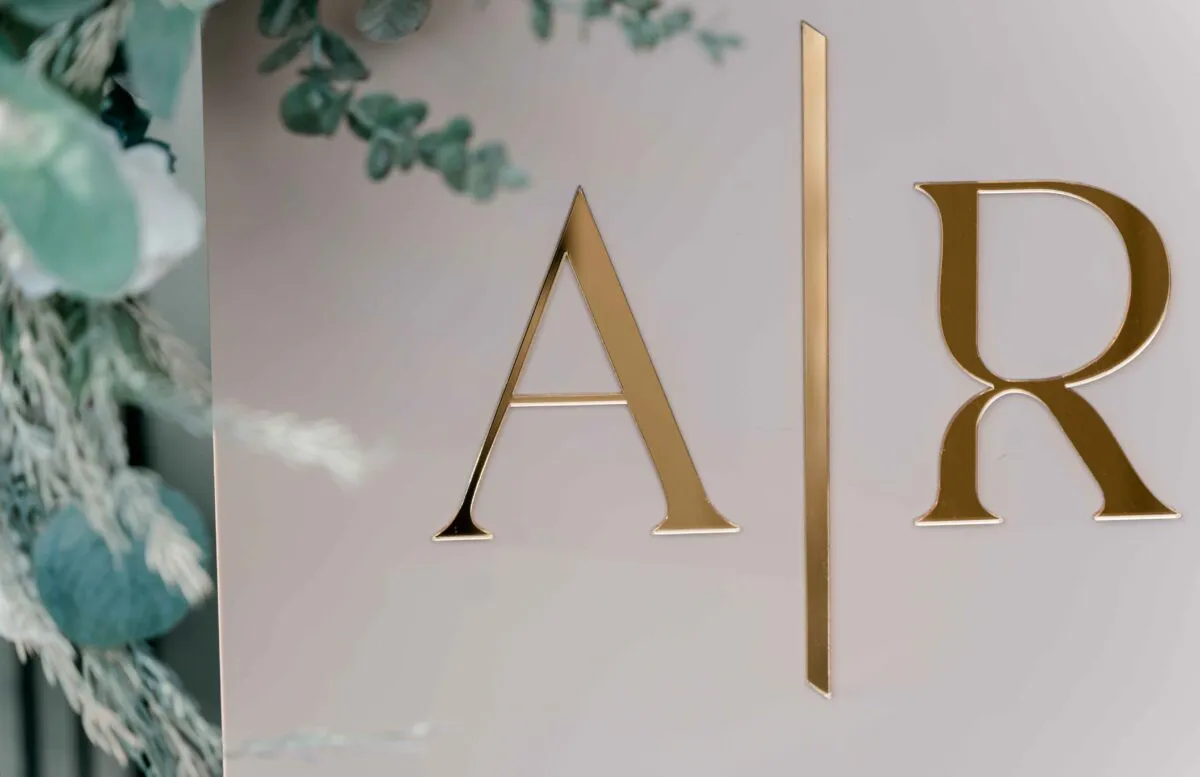 Close-up of a modern acrylic wedding sign featuring gold monogram lettering and a delicate eucalyptus floral arrangement framing the edge.