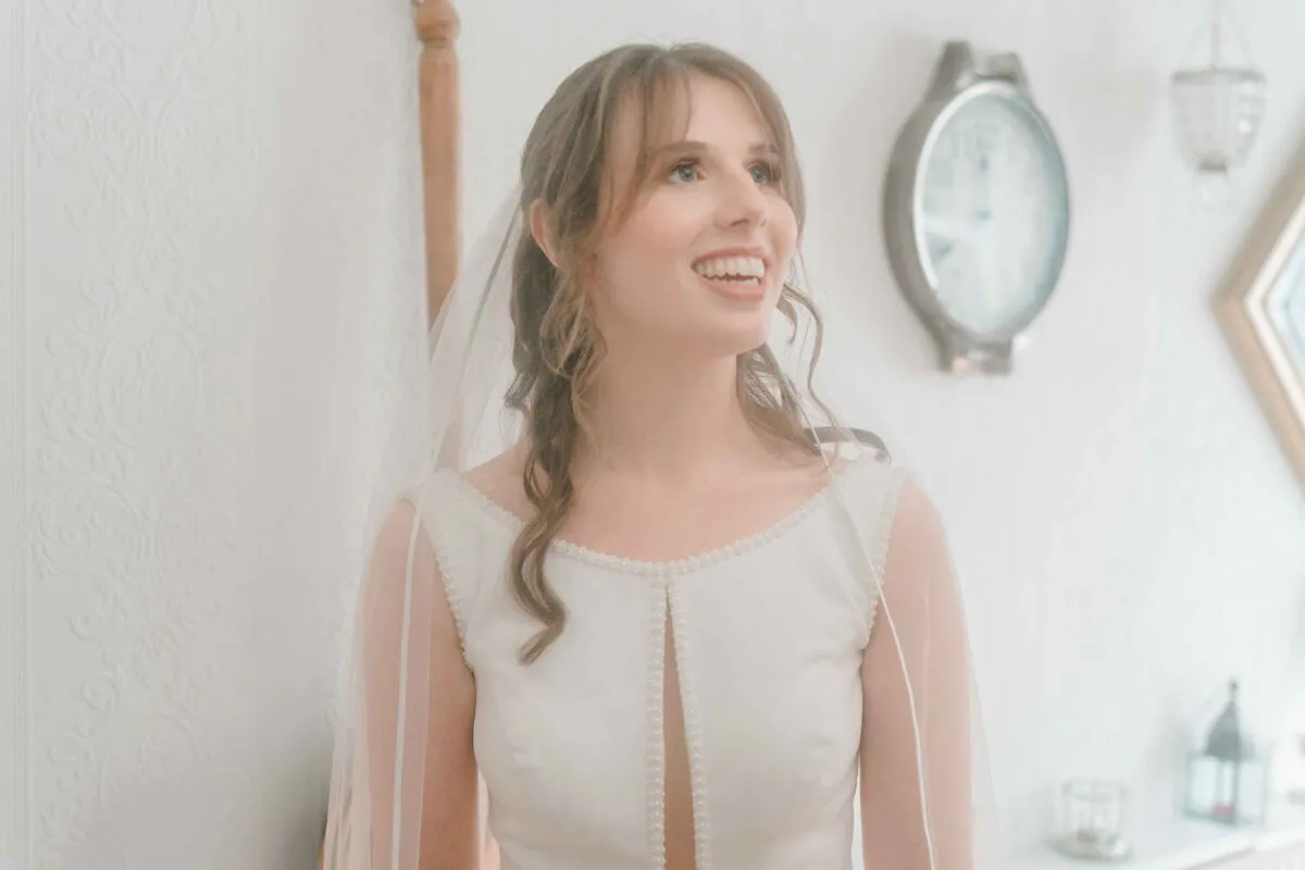 A joyful bride smiles brightly during her morning preparations, wearing a pearl-detailed dress and a delicate veil in a light-filled room.