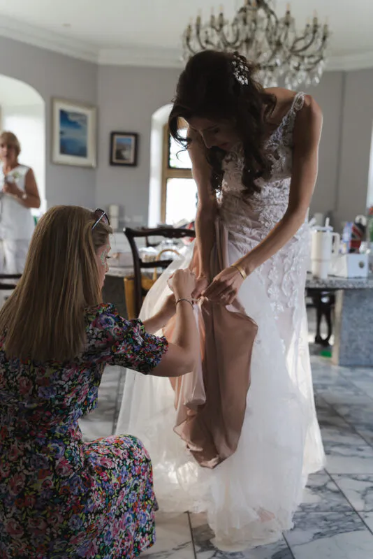 A bride in a detailed lace gown receives finishing touches on her dress from a bridesmaid in a floral dress during bridal preparations.