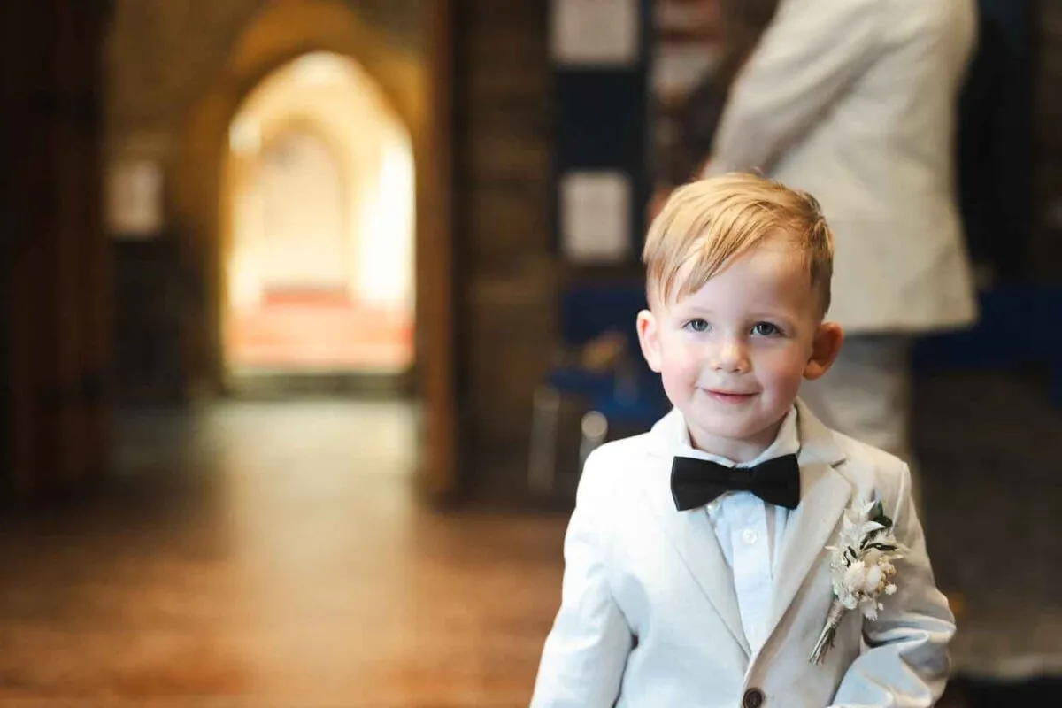 A young page boy wearing a smart cream suit, white shirt, black bow tie, and floral buttonhole smiles warmly inside a church.