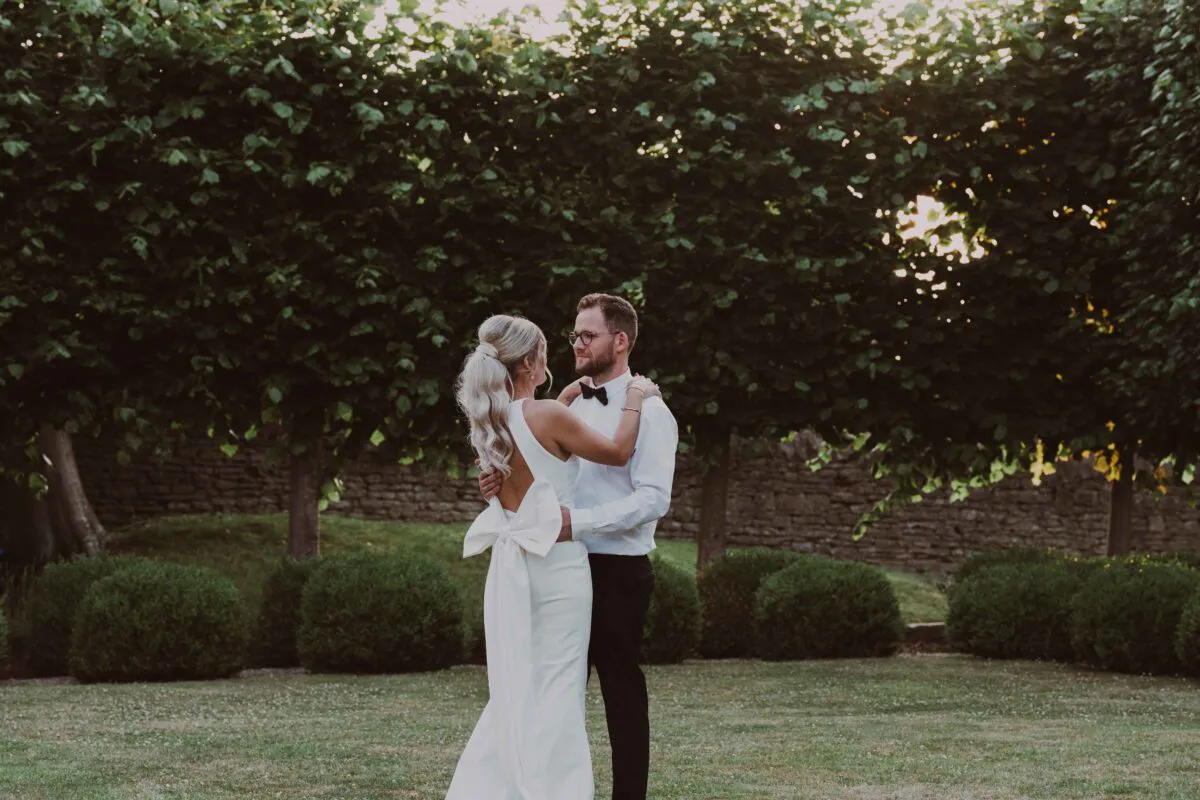 A bride and groom share a tender moment embracing outdoors in a manicured garden during golden hour, framed by lush green trees.