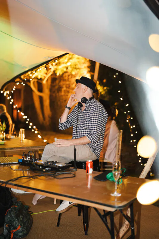 A wedding DJ wearing a fedora and checked shirt is focused at his mixing desk inside a marquee illuminated by glowing string lights.