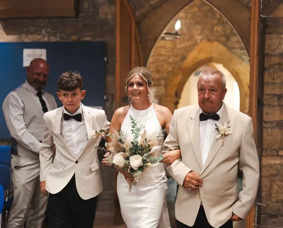 A smiling bride walks down the aisle of a stone church holding a hand-tied bouquet, escorted by her father and a young page boy.