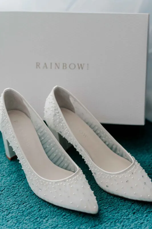 A pair of elegant white block-heel bridal shoes embellished with delicate pearls rests in front of their branded storage box.